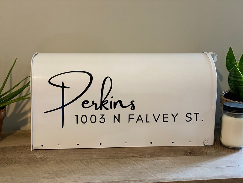 May include: A white mailbox displaying the name "Perkins" and address "1003 N Falvey St." in black script. The rectangular mailbox has a curved top and is on a wooden surface.