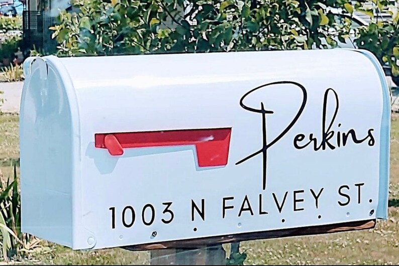 May include: White mailbox with a red flag and a black cursive "Perkins" name. Below the name is the address "1003 N FALVEY ST." The mailbox is mounted on a wooden post and is surrounded by greenery.