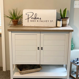 May include: White cabinet with two doors and a lower shelf. A white mailbox with the name "Perkins" and address is on top. Plants in pots and a candle sit on the cabinet. The cabinet has a light brown top and is on a carpeted floor.