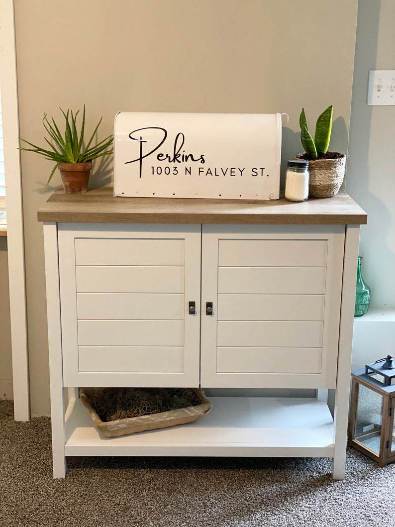 May include: White cabinet with two doors and a lower shelf. A white mailbox with the name "Perkins" and address is on top. Plants in pots and a candle sit on the cabinet. The cabinet has a light brown top and is on a carpeted floor.