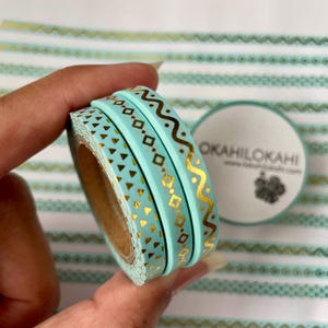 May include: A roll of pale blue washi tape featuring gold geometric designs, including triangles, diamonds, and wavy lines. The tape is partially unrolled, showing the inner cardboard core. A circular label with the text "OKAHILOKAHI" is visible.