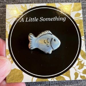 May include: A small, light blue fish-shaped pin with black details, set against a black circle with the text "A Little Something." The pin is on a card with a gold floral design.