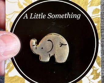 May include: A silver elephant pin with black detailing, displayed on a black circle with the text "A Little Something." The pin is presented on a card with a gold and white leaf design.