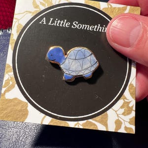 Könnte beinhalten: Ein blau-weißer Schildkröten-Ansteckpin mit goldenen Rändern wird auf einer Karte mit der Aufschrift "A Little Something" gezeigt. Der Pin ist ein kleines, dekoratives Accessoire. Die Karte hat einen schwarzen Kreis mit goldenen Blättern.