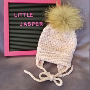 Könnte beinhalten: Eine rosa gestrickte Baby-Mütze mit einem weißen Fellbommel. Die Mütze hat Bänder, die unter dem Kinn gebunden werden können. Die Mütze liegt auf einer grauen Oberfläche mit einem rosa Buchstabenbrett, auf dem "LITTLE JASPER" steht.
