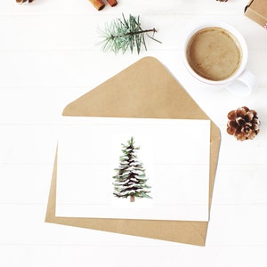 May include: A white card with a watercolor painting of a snow-covered pine tree. The card is sitting on a brown envelope on a white wooden surface. There is a cup of coffee, a pine cone, and a pair of scissors in the background.