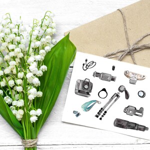 May include: A white card with a watercolor illustration of various photography equipment, including a camera, lens, tripod, and flash. The card is surrounded by a bouquet of white lily of the valley flowers.
