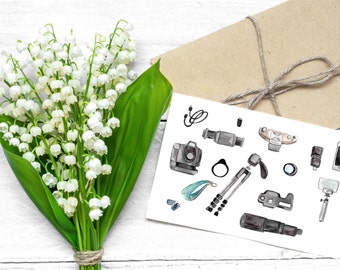 May include: A white card with a watercolor illustration of various photography equipment, including a camera, lens, tripod, and flash. The card is surrounded by a bouquet of white lily of the valley flowers.