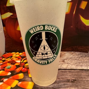 Puede incluir: Un vaso de plástico transparente con tapa y pajita, con un logotipo de "Weird Bucks Gravity Falls" en verde, negro y blanco. La taza está llena de purpurina y está colocada sobre un fondo temático de Halloween.