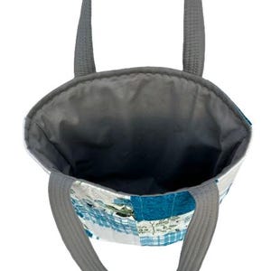 May include: A patchwork tote bag with gray handles and a gray interior. The exterior features a mix of blue, white, and green fabric squares. The bag is designed for carrying items.