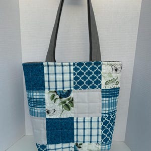 May include: A patchwork tote bag with gray handles. The bag features a variety of blue and white patterned squares, including floral, plaid, and geometric designs. The bag is held up by a person's hand.