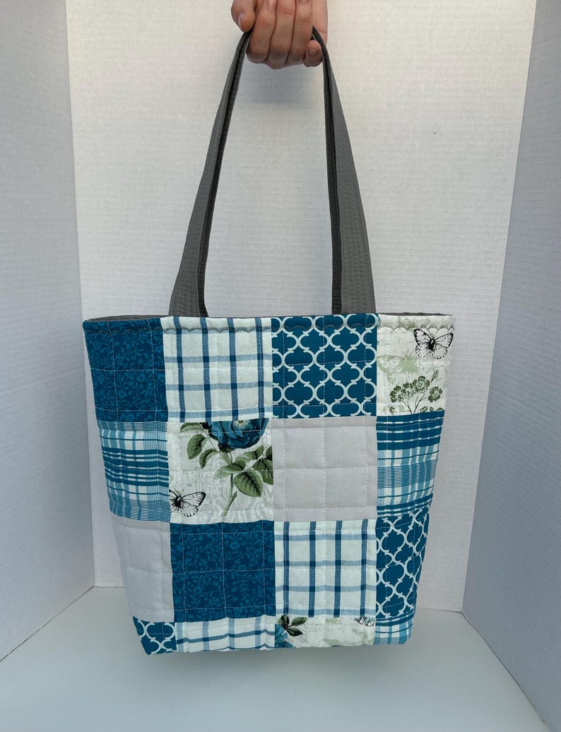 May include: A patchwork tote bag with gray handles. The bag features a variety of blue and white patterned squares, including floral, plaid, and geometric designs. The bag is held up by a person's hand.