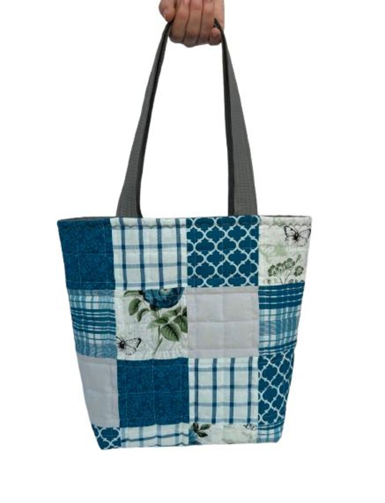 May include: A patchwork tote bag with gray handles. The bag features a variety of blue and white patterned squares, including floral, plaid, and geometric designs. Butterflies and flowers are also visible on some of the squares.