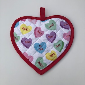 May include: Red heart-shaped pot holder with a white background and a quilted pattern. The pot holder is decorated with colorful candy hearts that have different messages on them, including "Love", "Sweet", "XOXO", "Hugs", "Kisses", and "Be Mine".