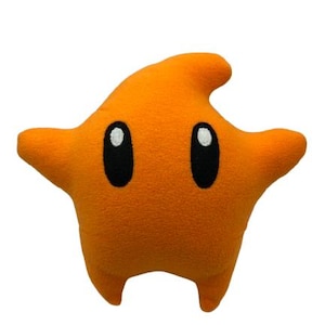 May include: An orange plush star-shaped cushion with black oval eyes and white highlights. The star has two arms, two legs, and a fin-like protrusion on top. The cushion is a solid orange colour and is set against a white background.