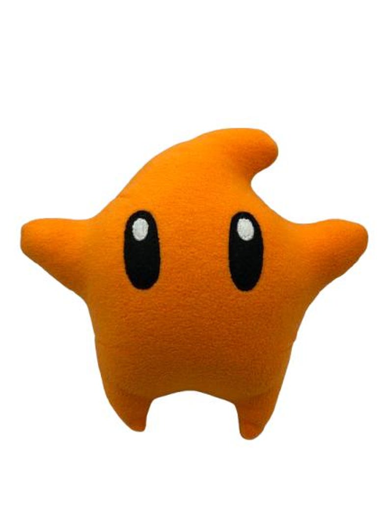 May include: An orange plush star-shaped cushion with black oval eyes and white highlights. The star has two arms, two legs, and a fin-like protrusion on top. The cushion is a solid orange colour and is set against a white background.