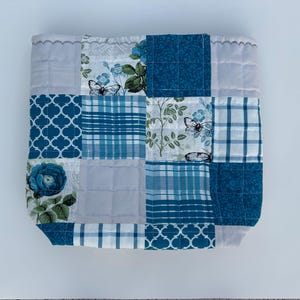 May include: A patchwork quilted square in shades of blue and white. The design incorporates floral, butterfly, and geometric patterns. The edges are finished with a scalloped trim, creating a decorative and handcrafted appearance.