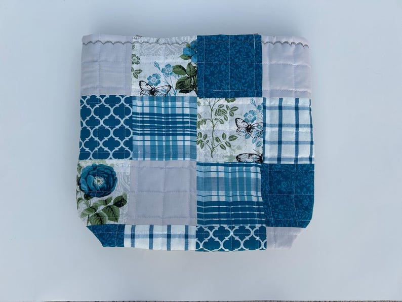 May include: A patchwork quilted square in shades of blue and white. The design incorporates floral, butterfly, and geometric patterns. The edges are finished with a scalloped trim, creating a decorative and handcrafted appearance.
