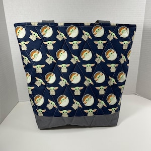 May include: A navy blue quilted tote bag featuring a repeating pattern of a cartoon character. The bag has gray accents at the base and handles. The bag is designed with a diamond pattern.