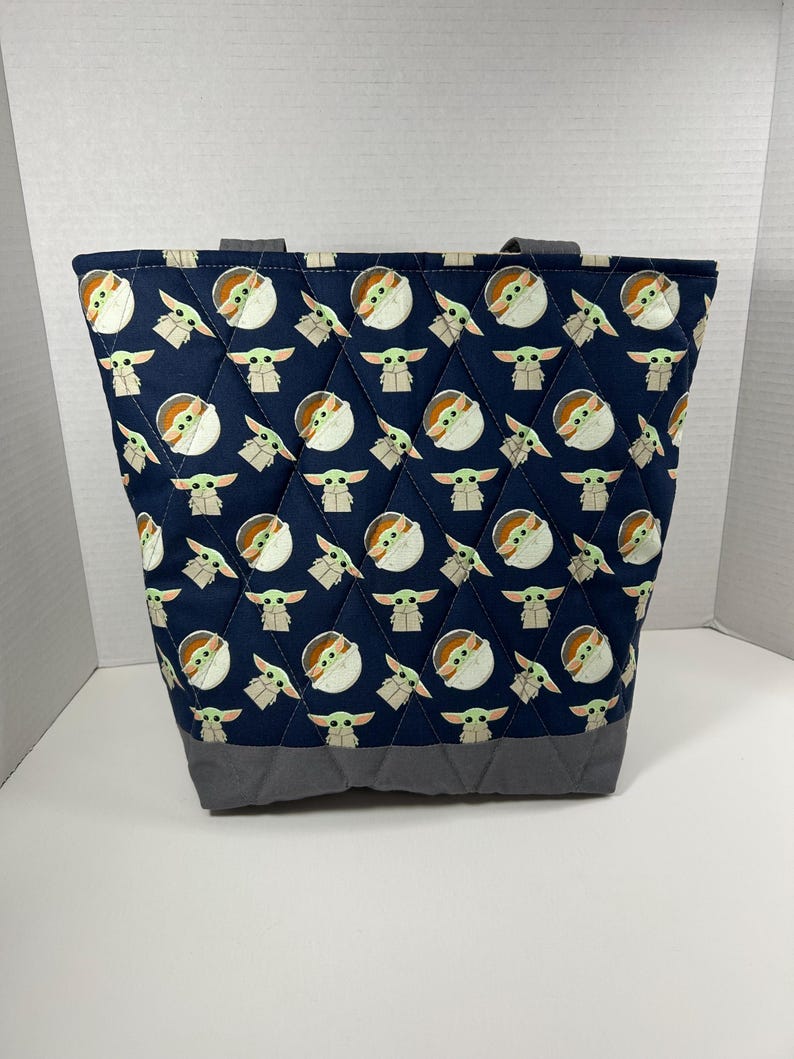 May include: A navy blue quilted tote bag featuring a repeating pattern of a cartoon character. The bag has gray accents at the base and handles. The bag is designed with a diamond pattern.
