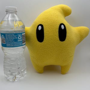May include: A yellow star-shaped plush toy with black oval eyes and a small mouth. The character has a curved top and two small legs. A clear plastic water bottle is next to the character.