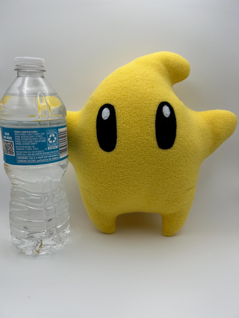 May include: A yellow star-shaped plush toy with black oval eyes and a small mouth. The character has a curved top and two small legs. A clear plastic water bottle is next to the character.