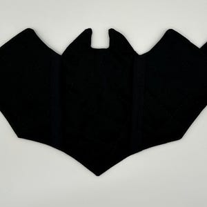 May include: A black quilted fabric oven mitt shaped like a bat with pointed ears and wide wings. The mitt features a small fabric loop on the upper right side for hanging. The quilted texture is visible across the surface of the black fabric.