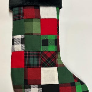 May include: Christmas stocking with a patchwork design. The stocking features a black faux fur cuff and a body composed of red, green, black, and white plaid squares. A black loop is attached to the top for hanging.