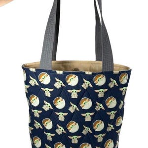 May include: A navy blue tote bag with a quilted pattern featuring a repeating design of a cartoon character. The bag has gray handles and a gray bottom. The interior lining is a tan color.