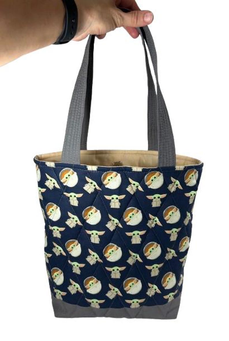 May include: A navy blue tote bag with a quilted pattern featuring a repeating design of a cartoon character. The bag has gray handles and a gray bottom. The interior lining is a tan color.