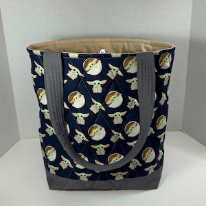 May include: A navy blue quilted tote bag featuring a repeating pattern of a green-skinned alien character. The bag has gray handles and a gray base. The interior lining is a light tan color.