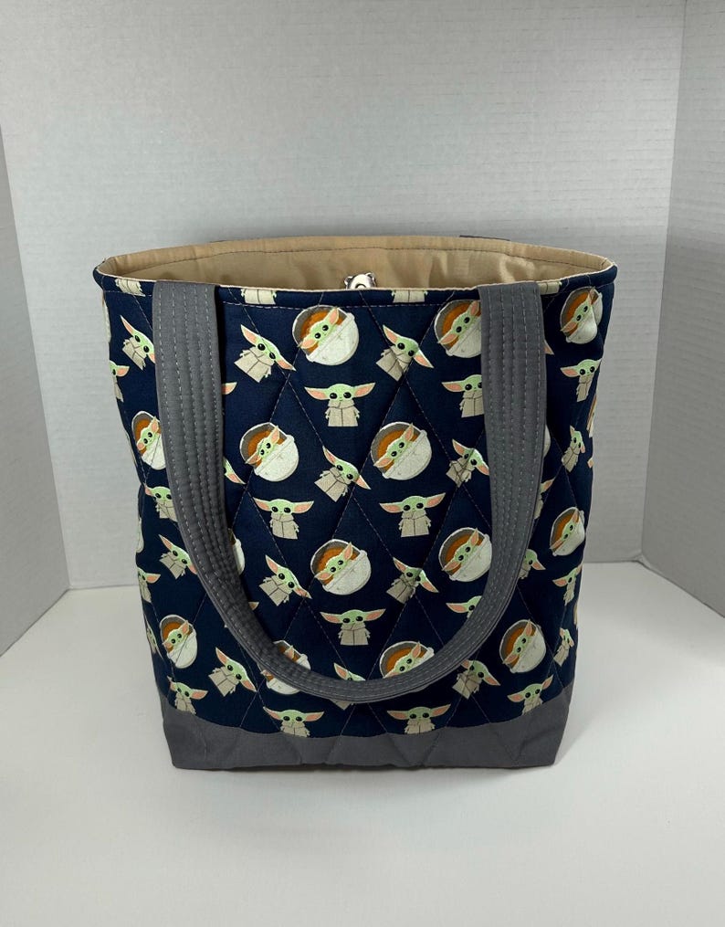 May include: A navy blue quilted tote bag featuring a repeating pattern of a green-skinned alien character. The bag has gray handles and a gray base. The interior lining is a light tan color.