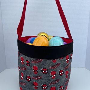 May include: A red and black fabric Easter basket with a red handle. The basket is filled with colorful plastic eggs and has a repeating pattern of Spider-Man's face and the word "Spider-Man".