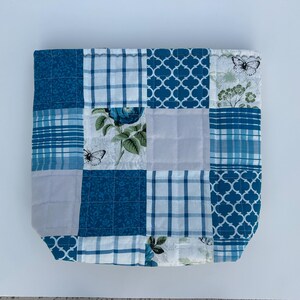 May include: A patchwork tote bag with a variety of blue and white patterns. The design includes floral prints, plaid, and geometric shapes. Butterflies and floral motifs are also present in the design.