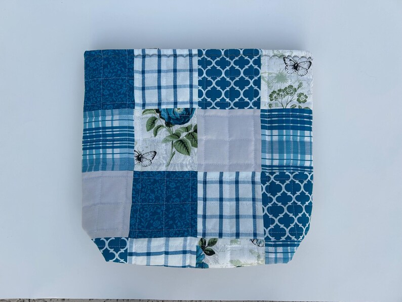 May include: A patchwork tote bag with a variety of blue and white patterns. The design includes floral prints, plaid, and geometric shapes. Butterflies and floral motifs are also present in the design.