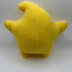 May include: A yellow, star-shaped cushion with a rounded top and two arms extending from the sides. The cushion has a soft, textured surface and small, rounded feet. The seams are visible down the centre.