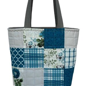 May include: A quilted tote bag with a patchwork design. The bag features squares of fabric in shades of blue, white, and gray, with floral, plaid, and butterfly patterns. It has gray handles and a gray top trim.