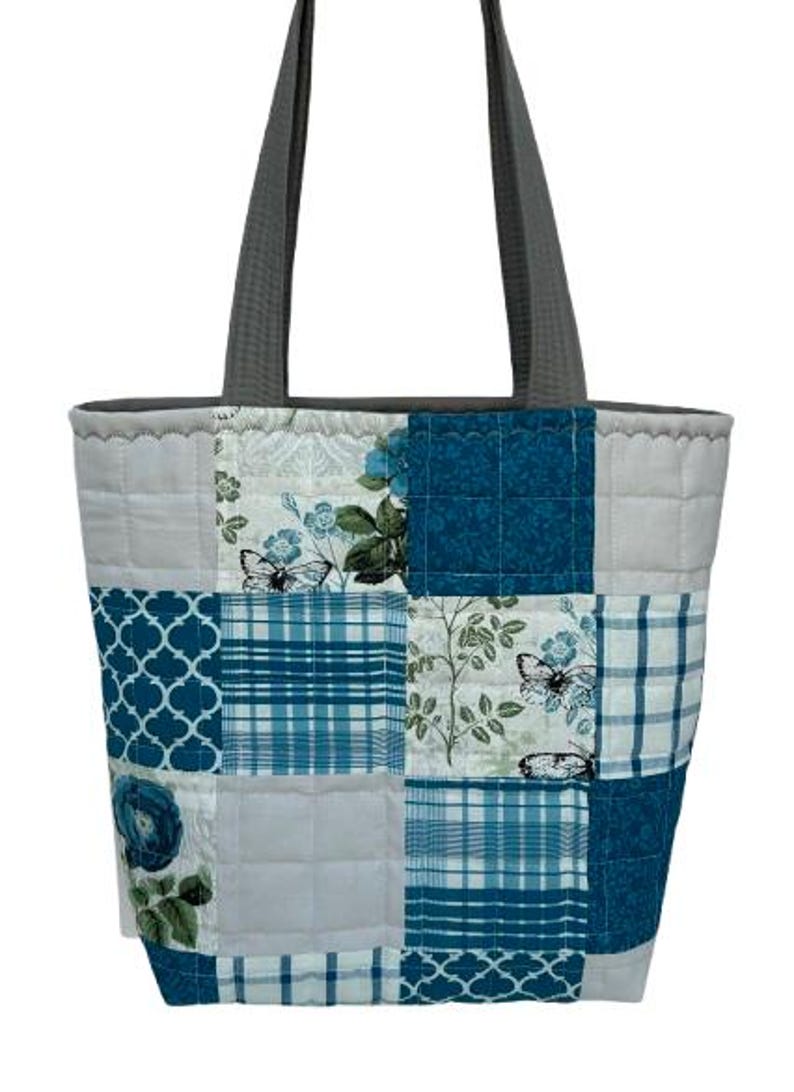 May include: A quilted tote bag with a patchwork design. The bag features squares of fabric in shades of blue, white, and gray, with floral, plaid, and butterfly patterns. It has gray handles and a gray top trim.