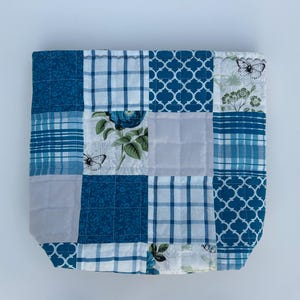 May include: A patchwork tote bag with a variety of blue and white patterns. The design includes floral, plaid, and geometric shapes. Butterflies are also featured in the design. The bag is square and appears to be quilted.