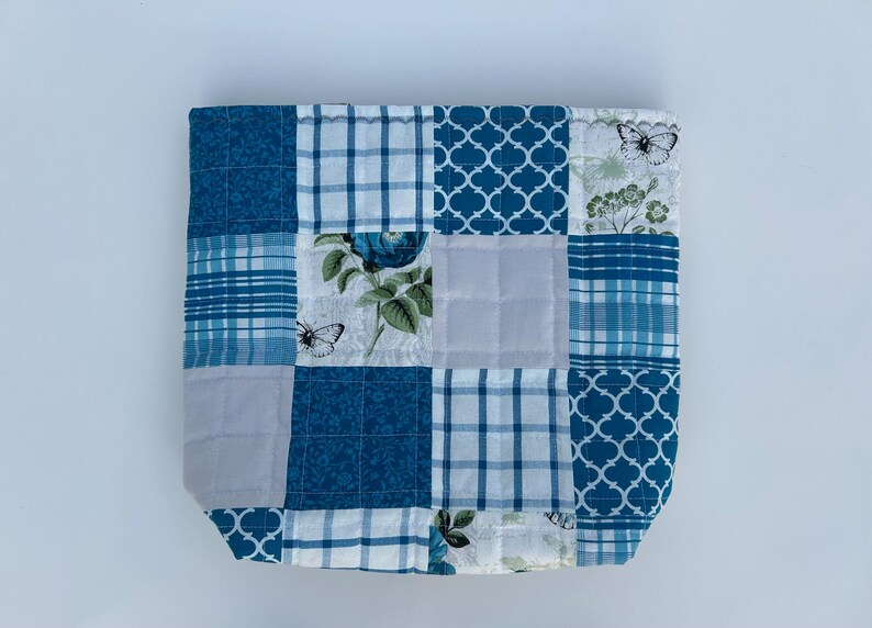 May include: A patchwork tote bag with a variety of blue and white patterns. The design includes floral, plaid, and geometric shapes. Butterflies are also featured in the design. The bag is square and appears to be quilted.