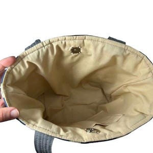 May include: An open tote bag with a beige interior and a small pocket. The exterior features a blue and white pattern. The bag has two handles and a magnetic closure. The bag is held open by a hand.