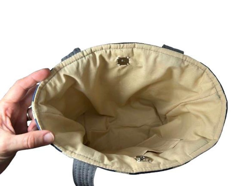 May include: An open tote bag with a beige interior and a small pocket. The exterior features a blue and white pattern. The bag has two handles and a magnetic closure. The bag is held open by a hand.