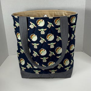 May include: Navy blue tote bag with a quilted pattern featuring a repeating design of a cartoon character. The bag has gray handles and a gray bottom. The interior lining is a light tan color.