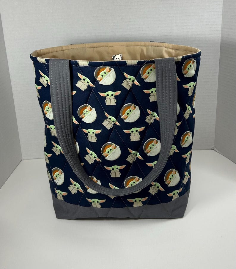 May include: Navy blue tote bag with a quilted pattern featuring a repeating design of a cartoon character. The bag has gray handles and a gray bottom. The interior lining is a light tan color.