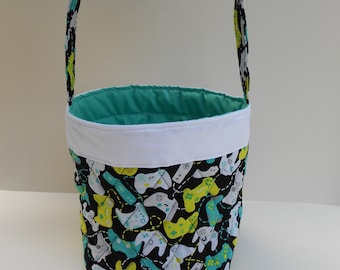 Peut inclure: Un panier de Pâques en tissu turquoise et blanc avec un imprimé noir et blanc de manettes de jeux vidéo. Le panier est doublé de tissu blanc et a une anse en tissu turquoise.