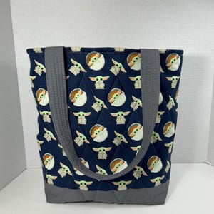 May include: Navy blue quilted tote bag featuring a repeating pattern of a cartoon character. The bag has gray handles and a gray base. The tote bag is designed with a diamond pattern.