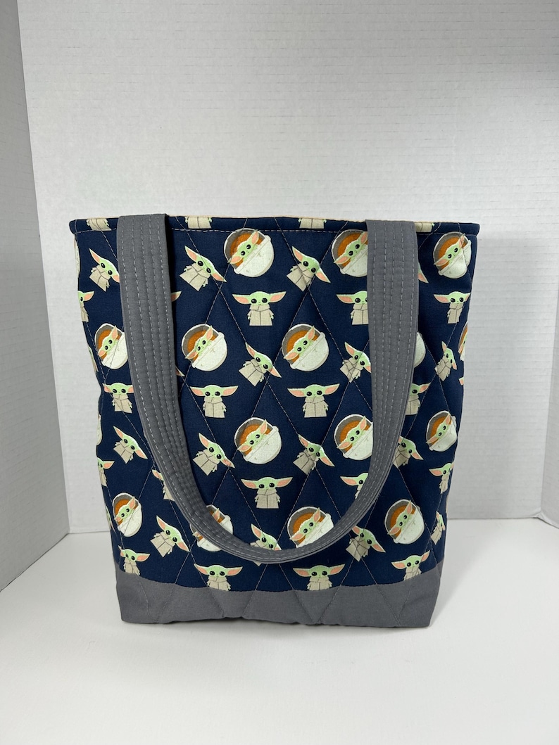 May include: Navy blue quilted tote bag featuring a repeating pattern of a cartoon character. The bag has gray handles and a gray base. The tote bag is designed with a diamond pattern.