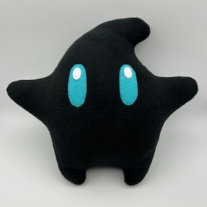 May include: A black star-shaped plush pillow with turquoise eyes and white highlights. The pillow has a rounded shape with two arms and two small feet. The plush toy is made of a soft material.
