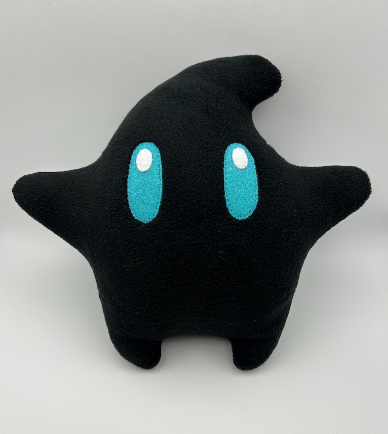 May include: A black star-shaped plush pillow with turquoise eyes and white highlights. The pillow has a rounded shape with two arms and two small feet. The plush toy is made of a soft material.