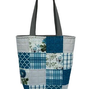 May include: A patchwork tote bag with a gray handle. The bag features a variety of blue and white patterned squares, including floral, plaid, and geometric designs. The bag is held up by a hand.